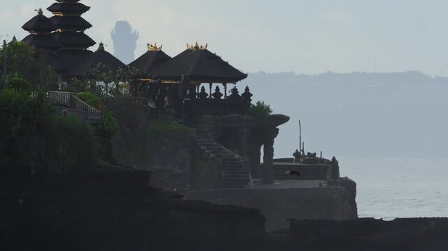 Silhouette Tanah Lot Temple Perched On Cliff Overlooking Ocean At Dusk, Multitiered Thatched Roofs, Stone Stairway, Small Figure Standing Near Edge, Misty Horizon, Gentle Waves Breaking Against Rocks,
