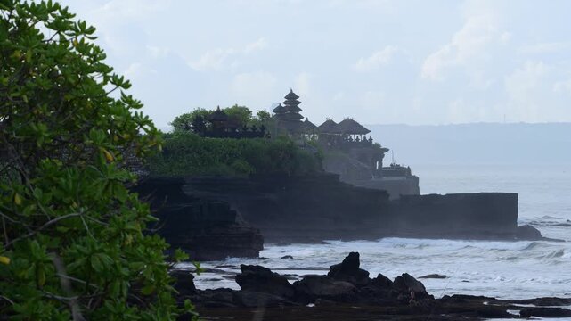 Coastal Temple Silhouette Tanah Lot Bali, Waves Crashing On Black Rocks With Lush Green Foreground, Ancient Pagoda Shapes On Cliff, Misty Horizon, Serene Worship Atmosphere And Dramatic Ocean Vista