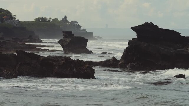 Rock Formation Framed By Surf,Towering Sea Stack With Pounding Waves,Mist Veil And Fierce Spray,Distant Temple Pagoda Tanah Lot On Cliff, Moody Light, Geology And Erosion Focus For Travel Documentary.