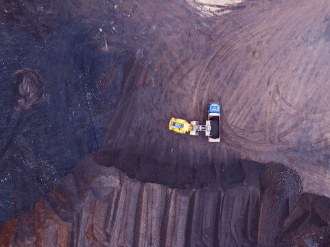 Aerial view of a yellow front-end loader and a blue dump truck working in a dark mining site with tire tracks on the ground in Iceland.