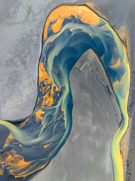 Aerial view of a braided river with vibrant blue and golden sediment patterns winding through the volcanic landscape of Iceland.
