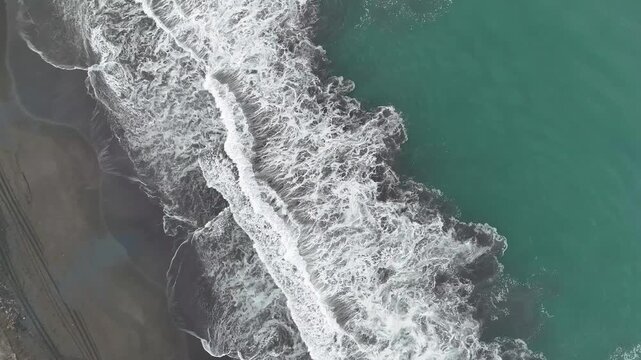 Stationary drone captures turquoise Pacific Ocean waves breaking onto a black sand beach from directly above, creating striking natural patterns along the tropical coastline.