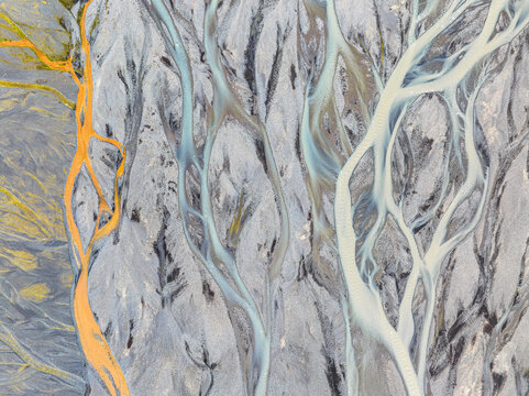 Aerial view of a complex braided river system with vibrant blue glacial meltwater and striking orange mineral deposits flowing through dark volcanic sediment in Iceland.