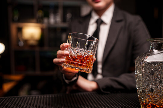 Woman bartender in a suit is handing a glass of whiskey across the bar counter.
