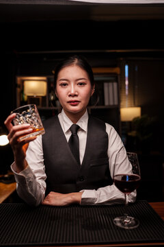 A professional female bartender holds a crystal glass containing red wine on the bar counter.
