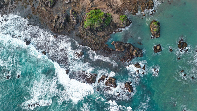 Aerial view of the rugged coastline with dark volcanic rocks, crashing turquoise waves, and lush green vegetation along the shore in Indonesia.