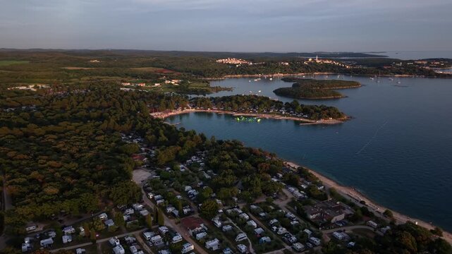 Dense rows of caravans wind through pine-shaded campground beside sheltered bay with floating water park, distant Vrsar's hilltop church catching golden light. Croatia. Aerial drone forward wide shot