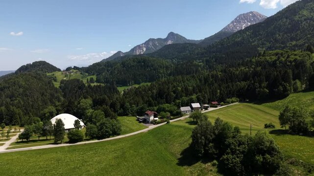 Traditional red-roofed Alpine farmhouse and white domed barn sit along curving gravel road, dwarfed by jagged limestone peaks rising above dense Austrian forest. Austria. Aerial drone view.