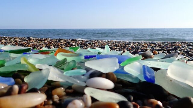 Colored sea glass with beach pebbles and shells in the mediterranean coast and in the background sea and waves with sunset and sky, sandy beach with coast glass, beach glass.