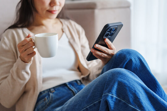 Young Asian woman drinking coffee and using mobile phone on a sofa at home in the morning