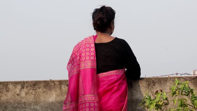 A Bengali woman with a hair bun rests her hands on a concrete roof railing outdoors. This scenery perfectly evokes sadness, solitude, and thinking emotions. Back view.