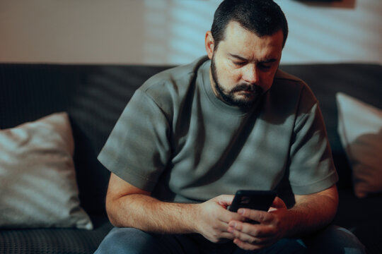 Sad Frowning Man Reading Urgent Message On Phone At Home. Anxious male character checks smartphone notification alone inside his quiet living room apartment 