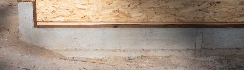 Closeup of poured concrete foundation with header wood boards anchored and OSB oriented strand board installed, dirt below, housing development project under construction  © knelson20