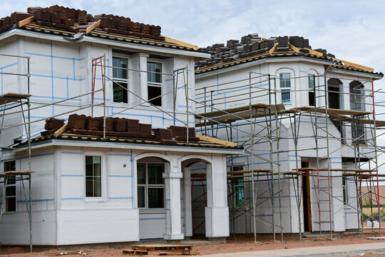 Two-story single family homes under construction with weather resistant building wrap, exterior scaffolding, and tile roofing in progress, representing residential development