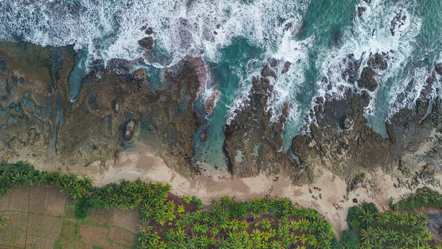 Aerial view of a tropical beach with rocky coastline, crashing waves, and lush palm trees bordering the sandy shore in Indonesia.