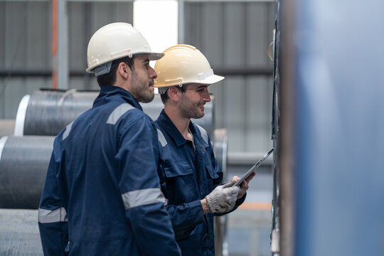 Two industrial engineers in hard hats and uniforms using digital tablet checking machine control panel in a metal sheet manufacturing factory
