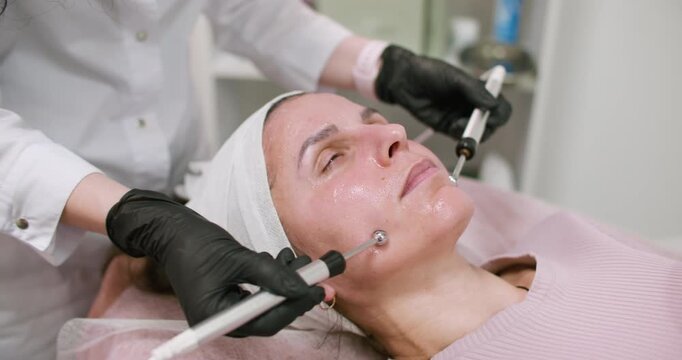 Microcurrent skincare. Female procedure. Esthetician in black gloves using professional modern device for lifting client face in salon.