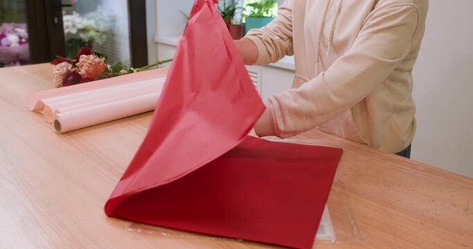 Floral workshop. Wrapping bouquet. Unrecognizable professional woman florist preparing red paper on table for chrysanthemum flowers.