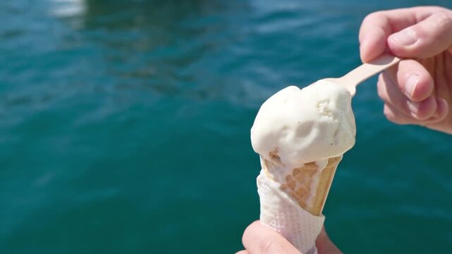 A person eats ice cream with a spoon. Only their hands are visible in the frame. The sea is visible in the background, creating a refreshing summer atmosphere.