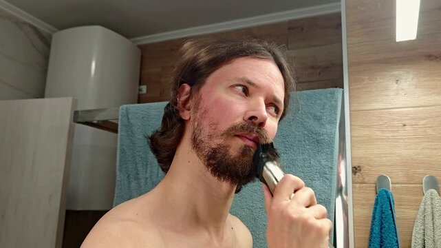 A man with long brown hair and a beard shaves with an electric razor in a bathroom. He appears focused and relaxed, surrounded by towels and wooden decor.