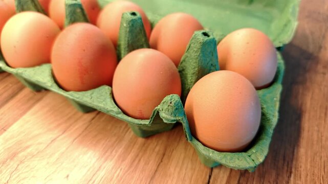 A close-up of brown eggs in a green cardboard tray on a wooden surface. The eggs are neatly arranged, showcasing their smooth texture and natural color.