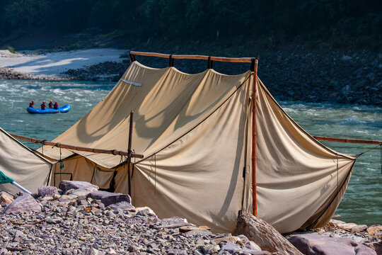 Camping tent on rocky riverbank and people rafting. Large safari tent pitched on stony riverbank with extreme rafting boat in background representing outdoor adventure tourism and camping lifestyle