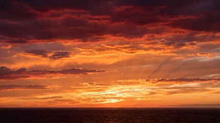 Fototapeta premium Dramatic fiery red and golden sunset sky over a dark ocean horizon. Breathtaking majestic clouds with glowing sun rays. Perfect nature landscape background for travel and weather concepts