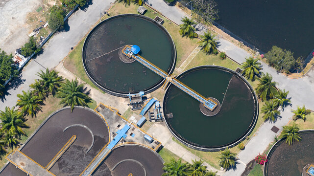 Aerial view of a wastewater treatment plant with circular sedimentation tanks surrounded by palm trees and asphalt paths in Phuket, Phuket, Thailand.