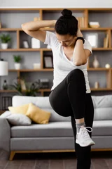 A young woman performs an abdominal crunch exercise while standing in her living room, focusing on her fitness routine © Sofiia Shunkina