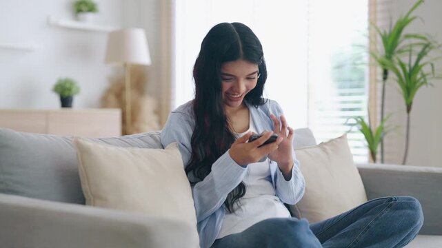Happy young asian woman relax on comfortable couch at home texting messaging on smartphone, smiling girl use cellphone, chatting online message, shopping online from home