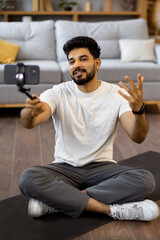 Obraz premium A young Indian man sits cross-legged on a yoga mat, holding a smartphone on a selfie stick to record a video