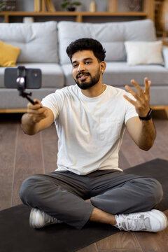 A young Indian man sits cross-legged on a yoga mat, holding a smartphone on a selfie stick to record a video