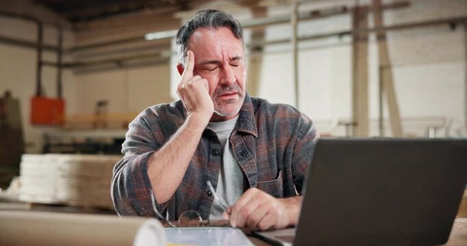 Headache, carpenter and laptop with man in workshop for fatigue, woodworking update and project crisis. Manufacturing, deadline and furniture supply chain with mature person in factory for inventory
