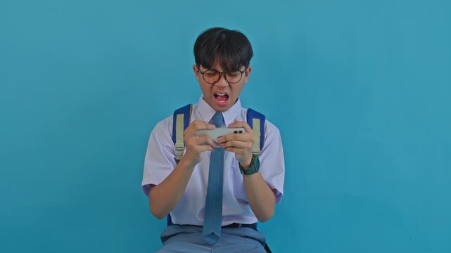 Frustrated schoolboy with backpack losing online phone game on blue background