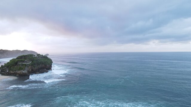 Aerial view of Pantai Drini, a rocky islet with lush vegetation surrounded by the Indian Ocean under a cloudy sky in Kecamatan Tanjungsari, Daerah Istimewa Yogya, Indonesia.