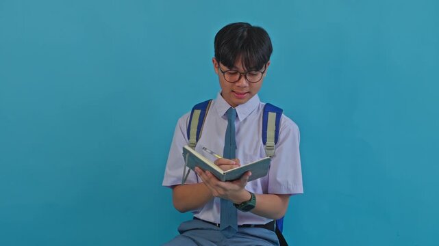 Schoolboy with backpack writing in notebook and getting bright idea on blue background