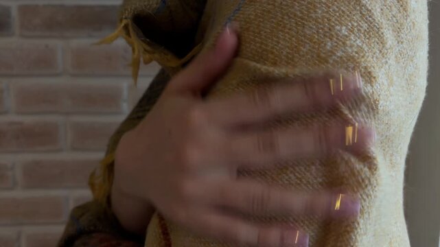 Woman with manicured nails wrapping a warm yellow scarf around her shoulders for comfort and coziness, feeling cold indoors on a chilly autumn day near a brick wall