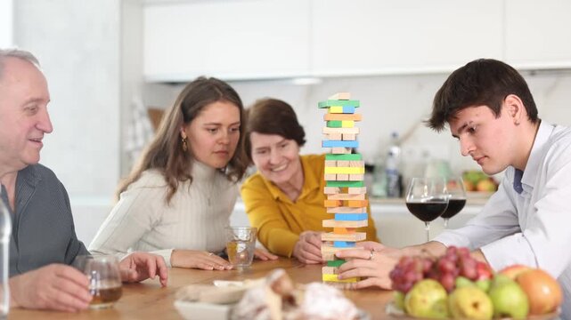 Happy young girl concentrating on removing block from tower while playing jenga, spending free time with boy-friend or husband and parents at light family home 