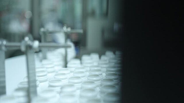 Rows of empty pill jars cluster on a pharma production workstation with blurred machinery and operator in the background.