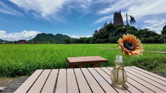 Single sunflower in a glass vase sits on a patio table facing Wat Tham Sua across a wide green countryside under a bright sky.