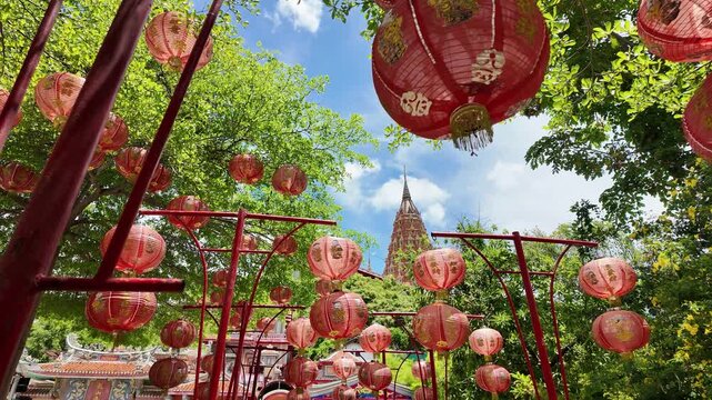 Rows of hanging red lanterns line a temple walkway under green trees with a Thai temple spire in the background on a sunny day.