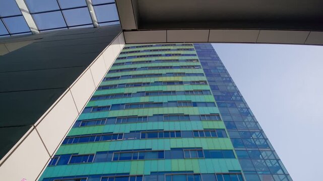 Looking up at a tall modern office building in Arnhem, Netherlands, featuring a distinctive green and blue geometric facade. Architectural structure frames the clear blue sky, emphasizing urban des...