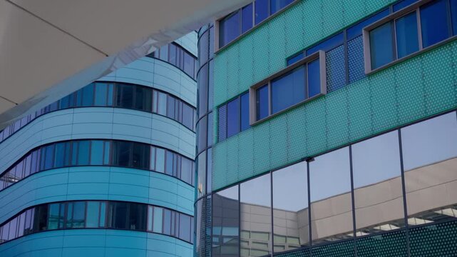 Close-up view of two distinct modern building facades displaying contrasting architectural styles. One features a curved blue surface, other has patterned turquoise panels and reflective glass wind...