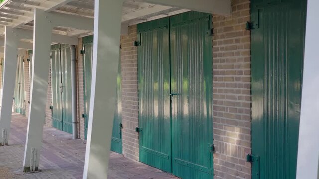 Row of historical brick buildings with striking green wooden doors and white support columns along a shaded outdoor walkway. Architecture suggests old utility structures or carriage houses.