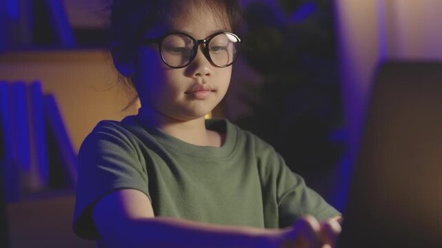 Young Boy with glasses working on a laptop in a darkened room with colorful ambient lighting, focused on screen to study or play video games