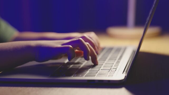 Close-up of hands typing on a laptop keyboard in a dimly lit room, with a blurred background and a cup of coffee on a wooden surface