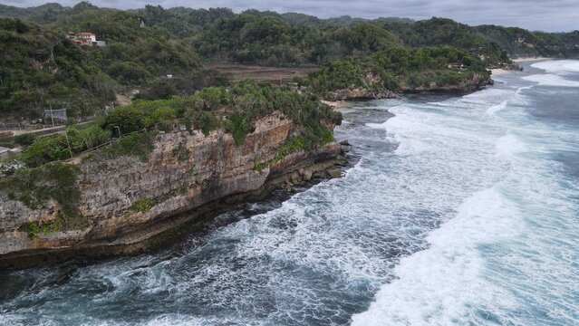 Aerial view of Indrayanti Beach with rugged limestone cliffs, crashing waves, and lush tropical vegetation along the coastline in Kecamatan Tepus, Daerah Istimewa Yogya, Indonesia.