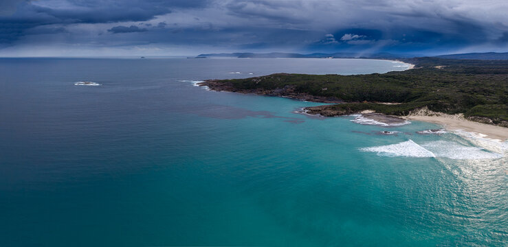 Aerial view of Conspicuous Cliff and the rugged coastline with turquoise waters, sandy beach, and dark stormy clouds overhead in Nornalup, Western Australia, Australia.