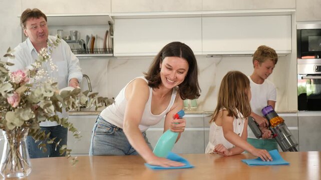 Positive woman washes kitchen table with rag with her daughter. Husband and son help to do cleaning in kitchen in background. Joint family house cleaning, household tasks, household help