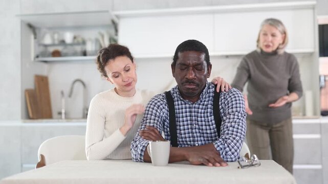 Middle-aged woman trying to calm angry man sitting at the kitchen table while mature woman quarreling to him standing behind . High quality 4k footage